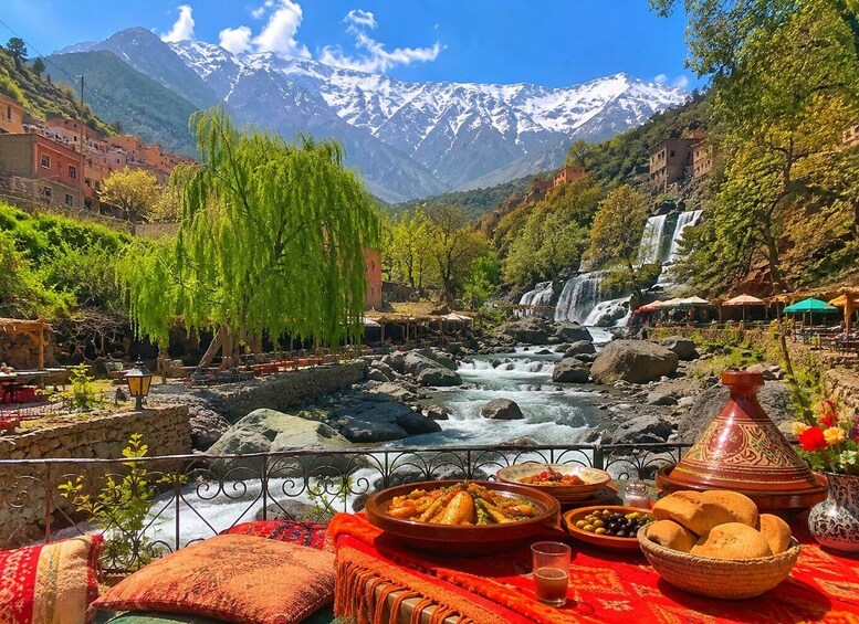 A riverside dining area with traditional dishes, a view of a waterfall, and snow-capped mountains in the background. 1