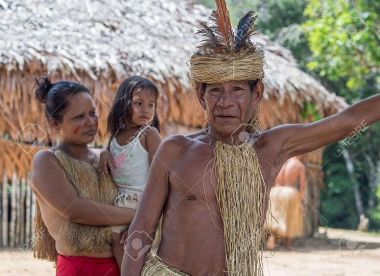 A person in traditional attire with a feathered headdress, standing outdoors with a thatched-roof structure in the background. 2