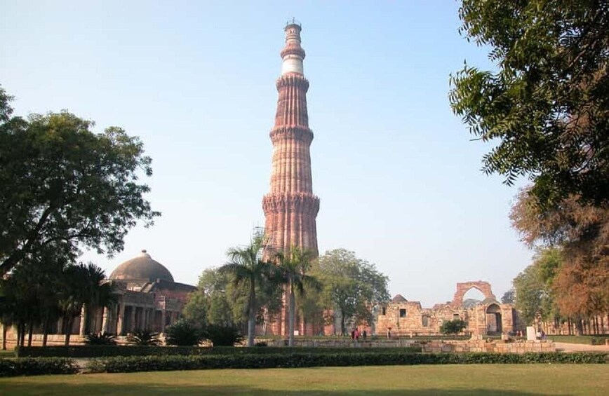 Qutub Minar on korkea, historiallinen torni, jonka ympärillä on puita ja muinaisia raunioita. 4