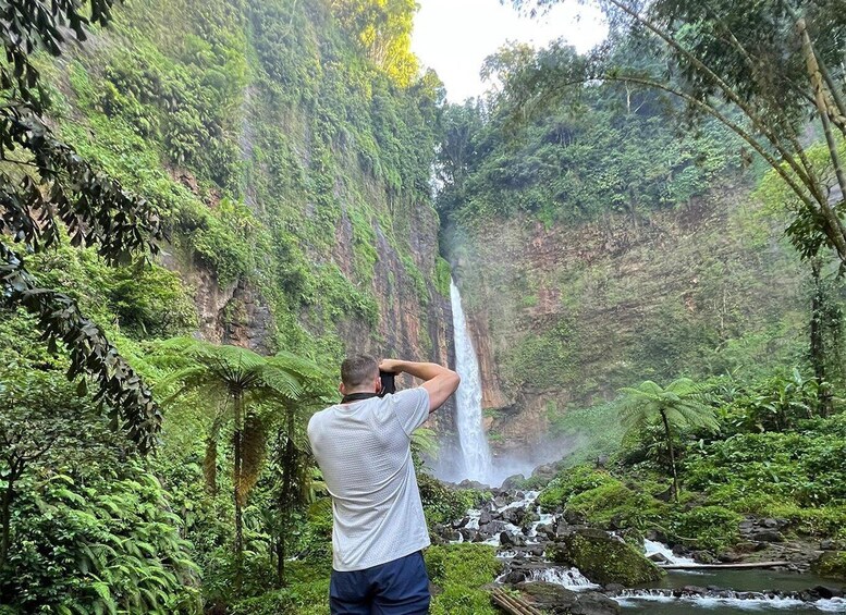 A person taking a photograph of a waterfall surrounded by lush greenery. 1