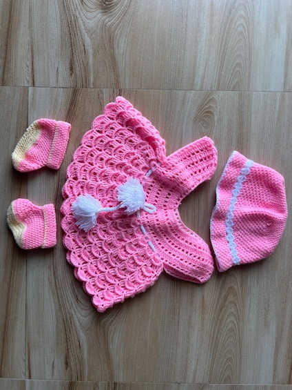 A pink crocheted baby outfit with matching mittens and a hat on a wooden floor. 3