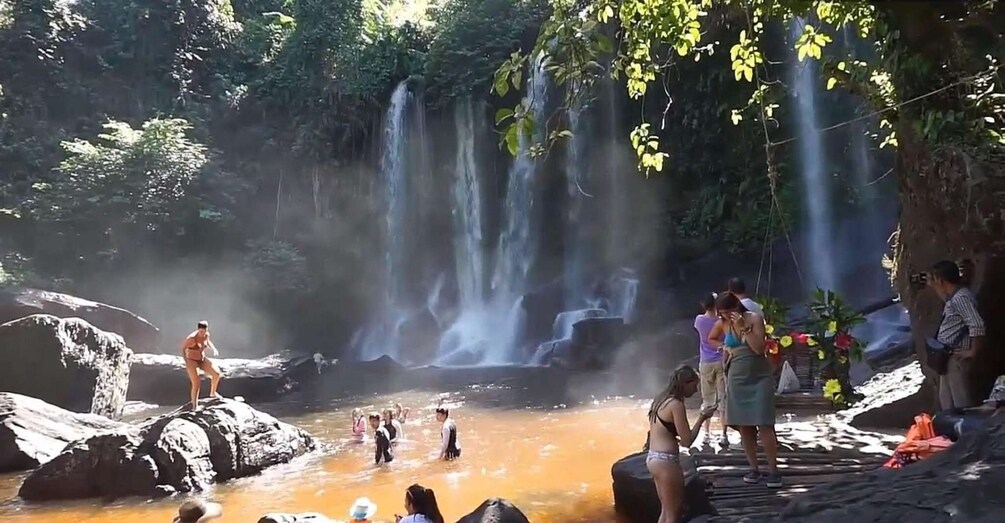 A waterfall in a forested area with people standing and sitting around a small pool of water. 1