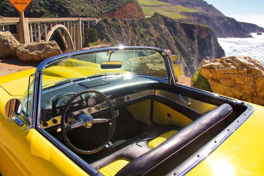 A yellow convertible car with a black interior, parked near a coastal cliff with a bridge in the background. 4