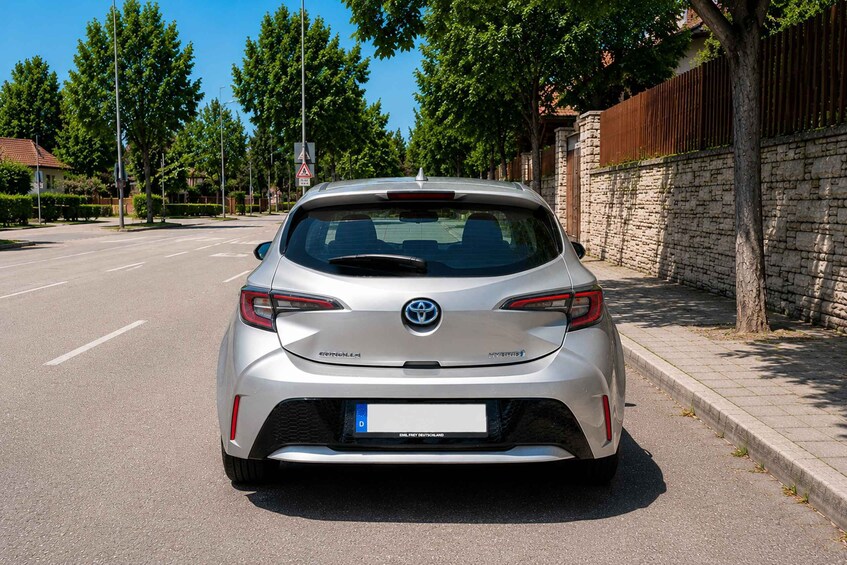 A silver Toyota C-HR parked on a residential street with trees and a stone wall in the background. 3