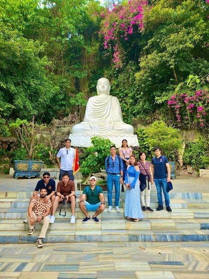 A group of people posing in front of a large Buddha statue surrounded by lush greenery and pink flowers. 5
