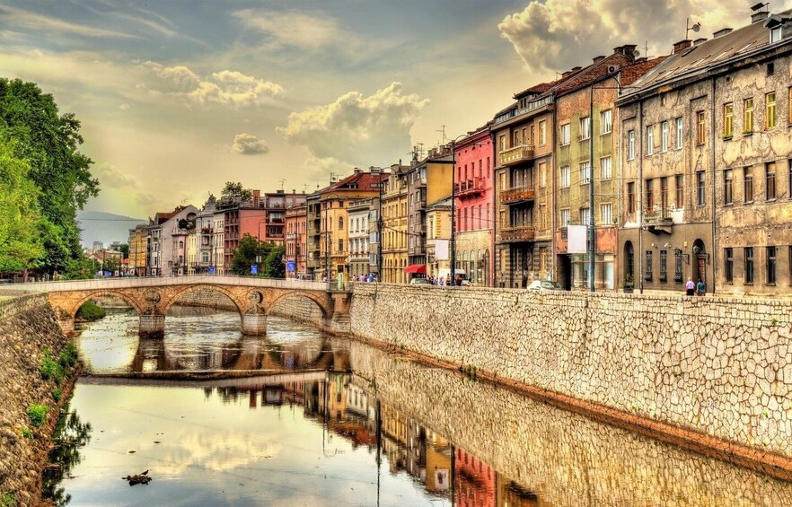 A stone bridge over a calm river, reflecting colorful buildings and a cloudy sky. 1