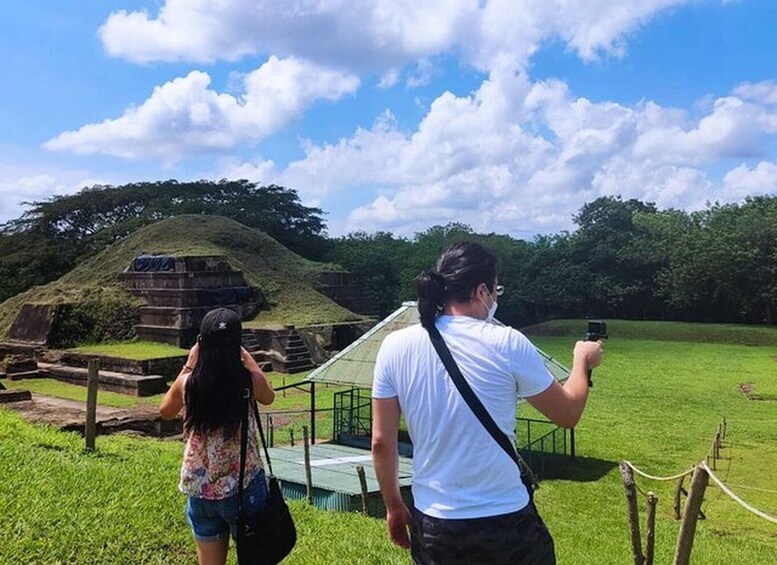 Two people taking a photo of an ancient pyramid-like structure surrounded by grass and trees. 3