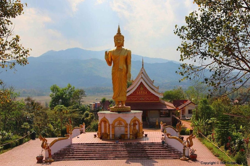 A large golden Buddha statue on a pedestal with a temple in the background and mountains in the distance. 1