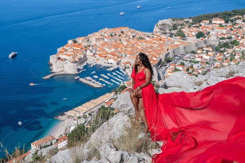 A woman in a red dress standing on a rocky outcrop overlooking a coastal town with a historic fortification and a marina. 1