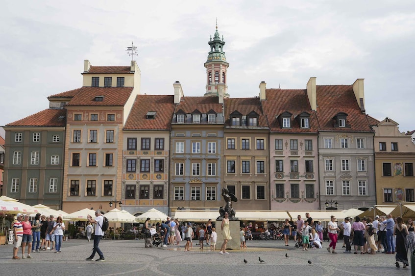 A bustling square with people walking, a statue, and historic buildings with red-tiled roofs. 1