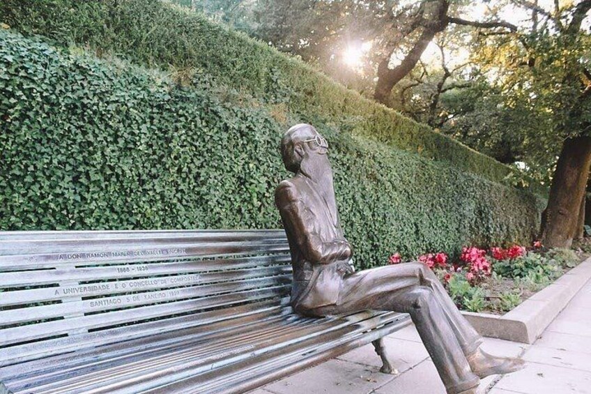 Una estatua de bronce de una figura sentada en un banco, con inscripciones, rodeada de follaje y flores. 2