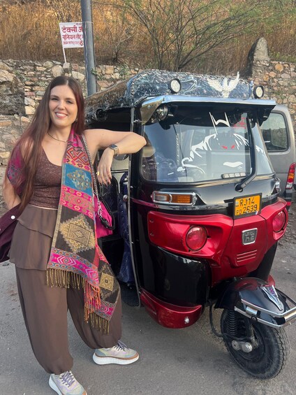 A person standing next to a red auto-rickshaw with a license plate, a stone wall, and dry trees in the background. 5