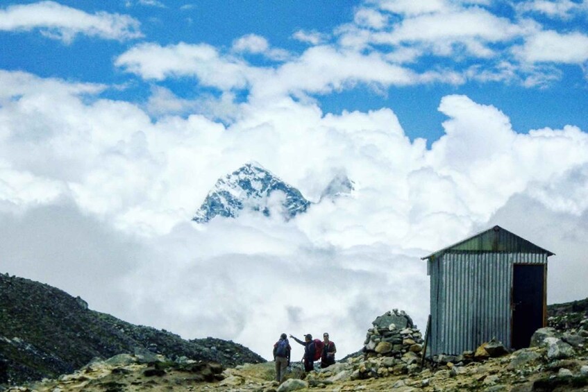 Un pico montañoso con nubes abajo, terreno rocoso y un pequeño refugio metálico. 4