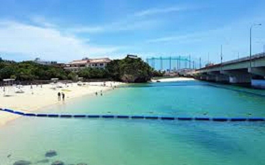 A beach with clear turquoise water, a sandy shore, and a bridge in the background. 1