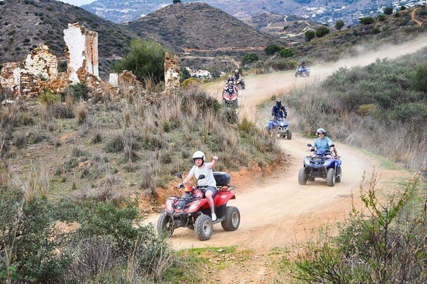 Eine Gruppe von Personen fährt mit Quads auf einer unbefestigten Straße, im Hintergrund sind Ruinen zu sehen. 1