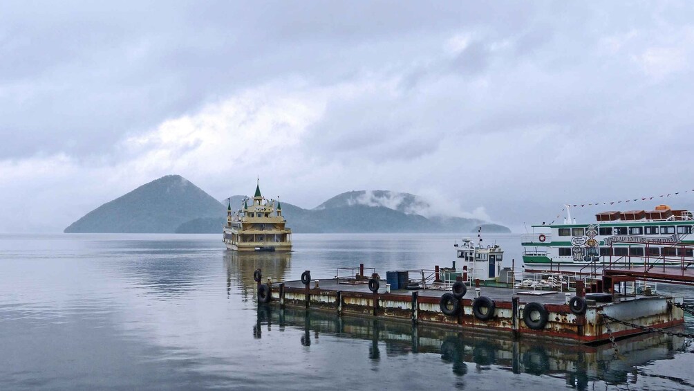 A calm lake with a large boat docked at a pier, a smaller boat in the water, and mountains in the background. 3