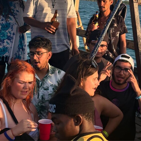 A group of people on a wooden pier by the water, holding drinks and engaging in conversation. 3