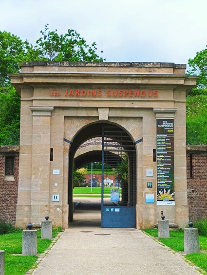 An entrance to Les Jardins Suspendus with an archway and a signboard listing events. 2