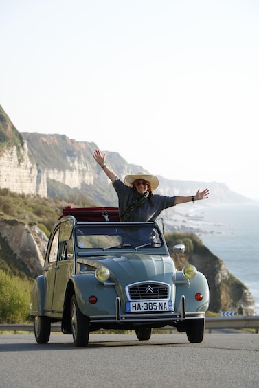 A vintage car with a person waving, driving on a coastal road with cliffs in the background. 1