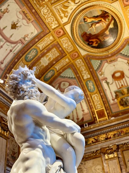 A marble statue of a seated figure with a crown of laurel wreaths, set against a richly decorated ceiling with intricate patterns and a central circular medallion. 1
