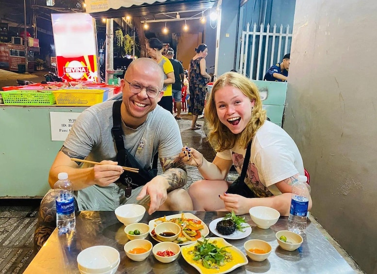 Two people dining at an outdoor food stall with various dishes and condiments in front of them. 1
