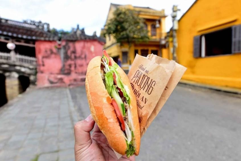A hand holding a sandwich with a paper bag labeled "PHUONG" and a historic building in the background. 5