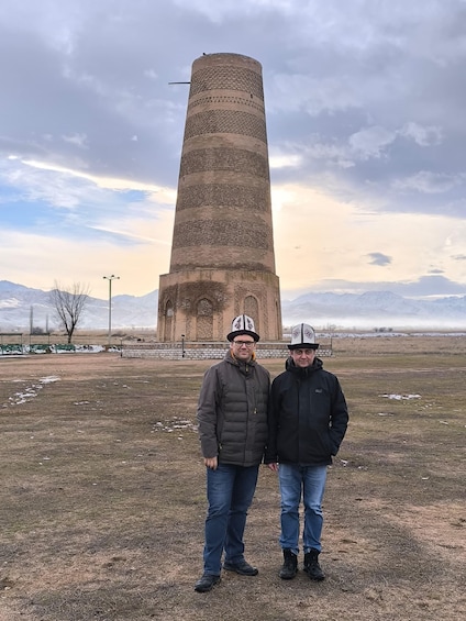 Two people standing in a field with a tall, patterned tower in the background. 2