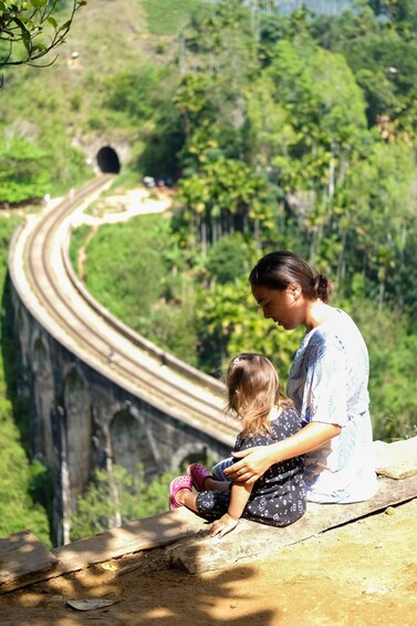 A woman and a child sitting on a ledge overlooking a winding road and a tunnel in a lush green landscape. 2