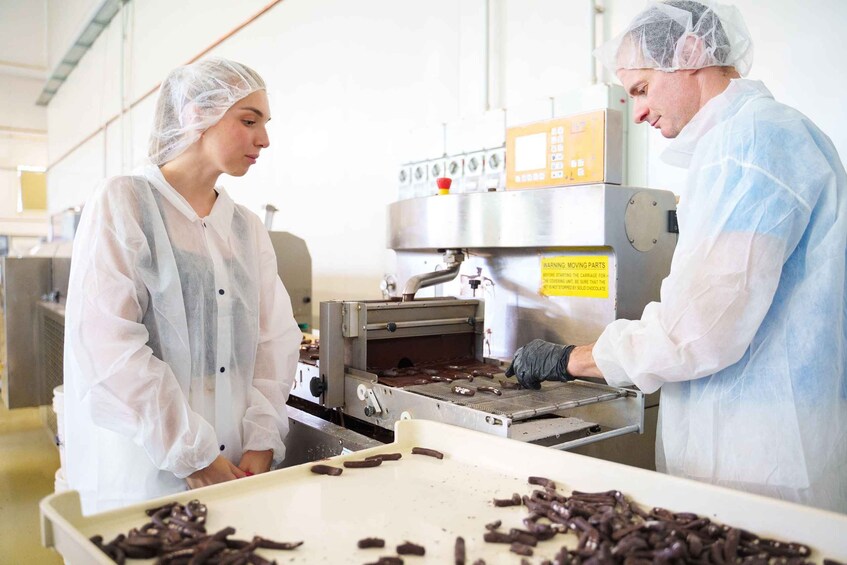 Two people in a factory setting, wearing protective clothing, handling chocolate pieces on a conveyor belt. 2