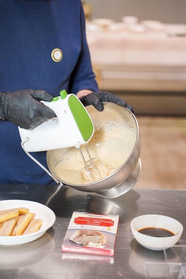 A person in a blue uniform and black gloves mixing batter in a metal bowl using an electric mixer. There are plates with pastries and a packet of cake mix on the table. 3