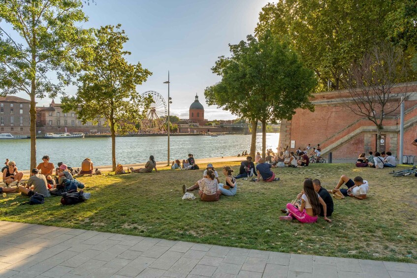 People sitting on grass by a river with a Ferris wheel and a domed building in the background. 2