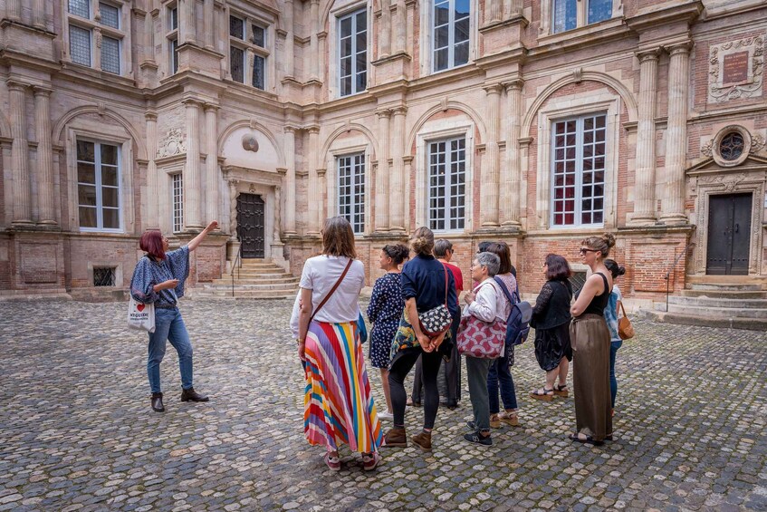 A group of people standing in a cobblestone courtyard in front of a historic building with large windows and columns. 1
