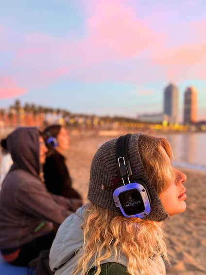 A person wearing headphones with a visible logo, sitting on a beach with a city skyline in the background. 1
