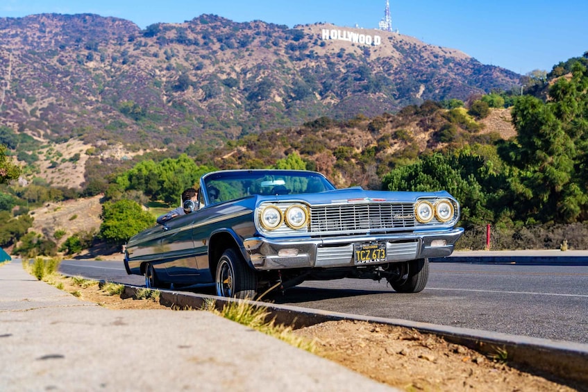 A classic blue car parked on a road with the Hollywood sign in the background. 1