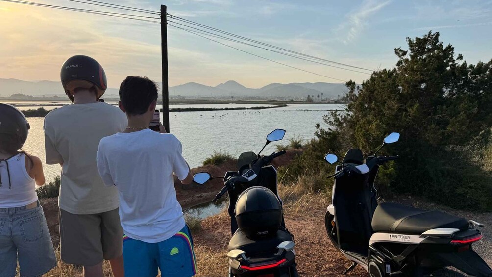 Three people standing by a body of water with mountains in the background and two scooters parked nearby. 2