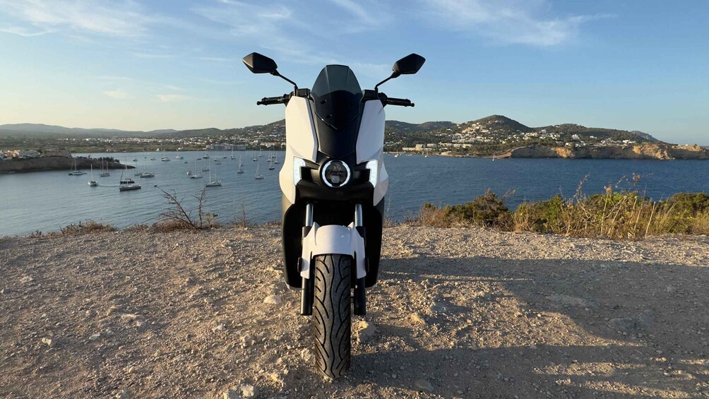 A white motorcycle parked on a rocky shore with a view of a coastal town and boats in the background. 5