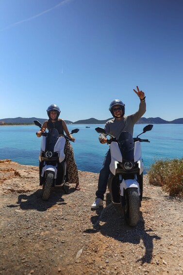 Two people on scooters by a lake with mountains in the background. 1