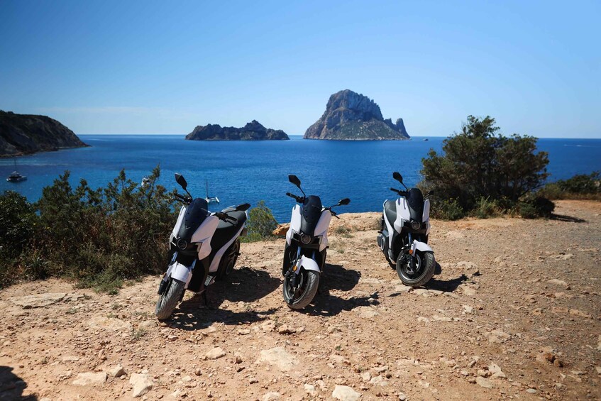 Three scooters parked on a rocky path with a view of the sea and a prominent rock formation in the distance. 4