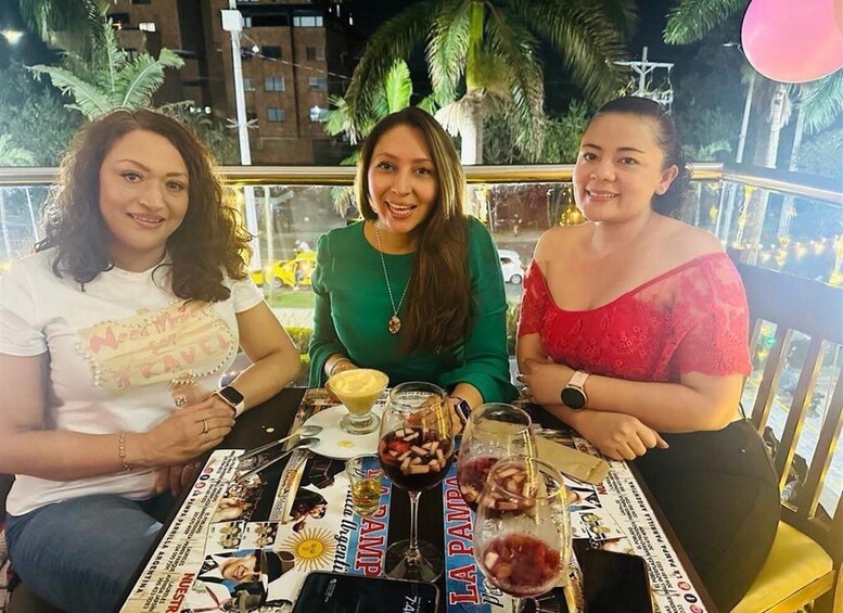 Three people sitting at a table with drinks and food, outdoors with palm trees in the background. 2