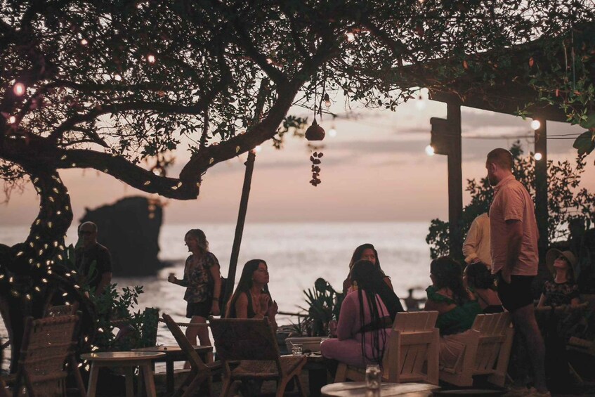 A group of people seated at tables under a tree with hanging lights, overlooking a body of water at sunset. 2