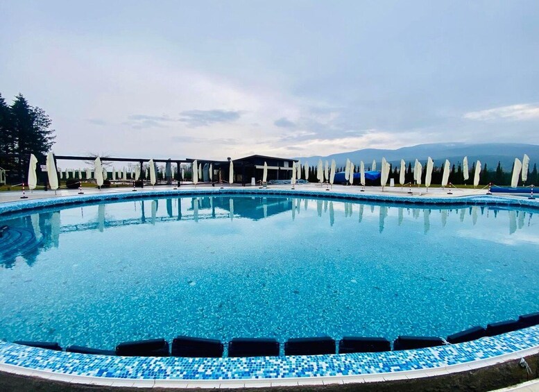 A large, clear blue pool with tiled edges, surrounded by white umbrellas and a building in the background. 3