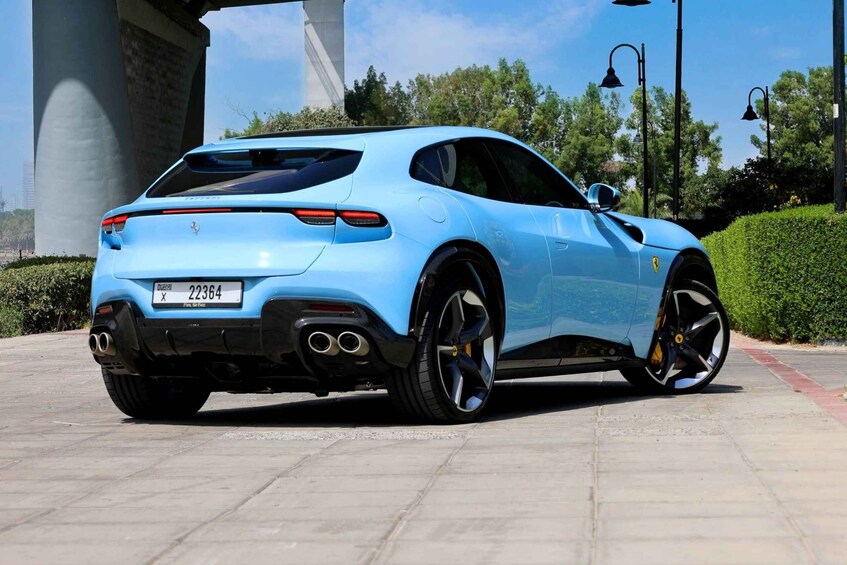 A light blue sports car parked on a paved surface with a large concrete structure and greenery in the background. 2