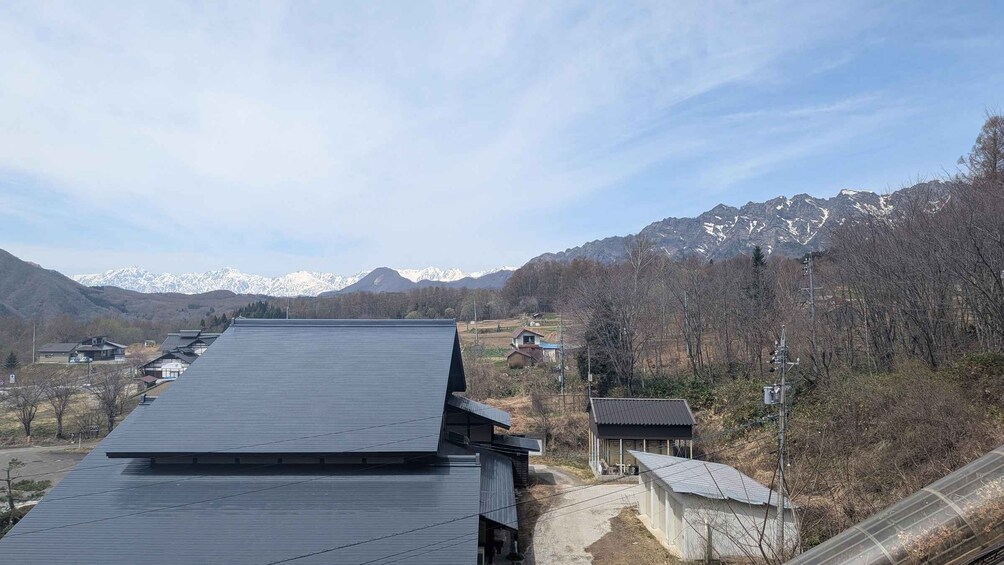 A rural landscape with a long building with a dark roof, surrounded by trees and mountains in the background. 3