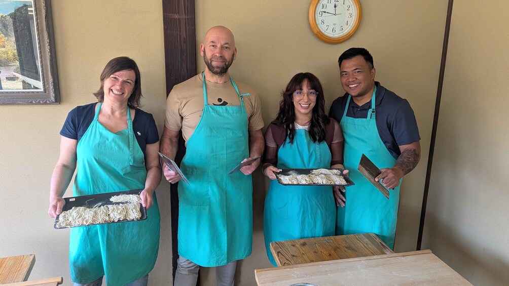 Four people in aprons holding trays of food, standing in a room with a clock on the wall. 1