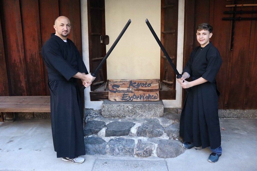 Two individuals in black robes holding swords in front of a wooden door with a sign that reads "Kyoto Samurai Experience". 1