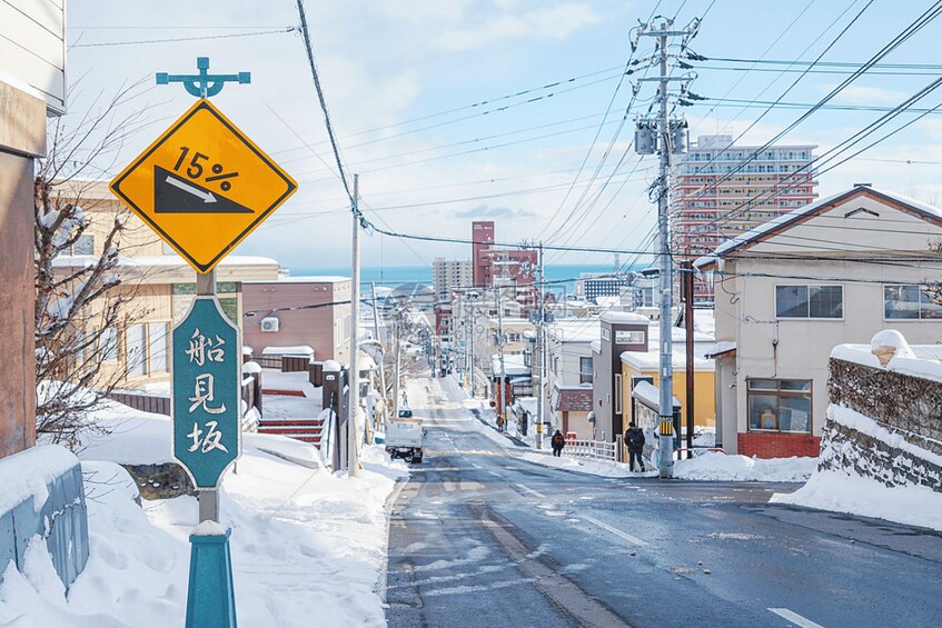 A snowy urban street with a 15% grade warning sign, buildings, and power lines. 5