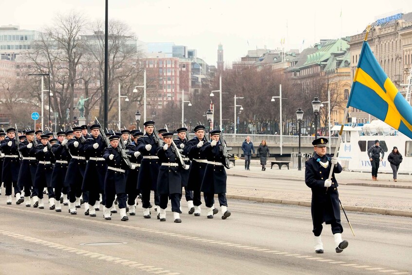 Sotilasparaati, jossa univormuihin pukeutuneet sotilaat marssivat rivissä, Ruotsin lippu ja kaupunkikuva, jossa taustalla näkyy rakennuksia ja silta. 4
