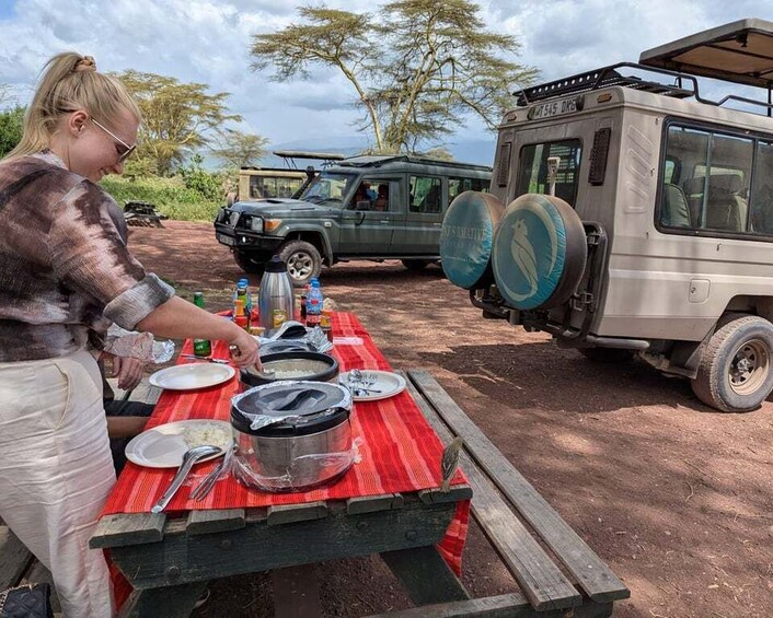 A person preparing food at a picnic table with a vehicle and safari equipment in the background. 1