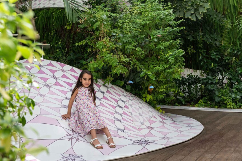 A child sitting on a curved, patterned surface surrounded by lush greenery. 2