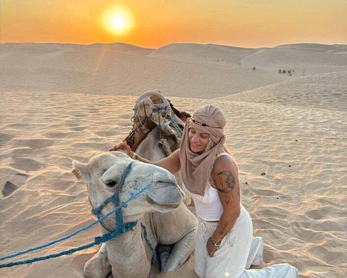 Une personne assise sur un chameau dans un désert, avec des dunes de sable et un coucher de soleil en arrière-plan. 4